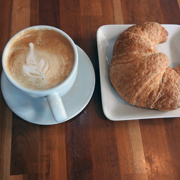 A latte with foam art and a croissant on white plates, on a wooden table at Cafe La Taza.