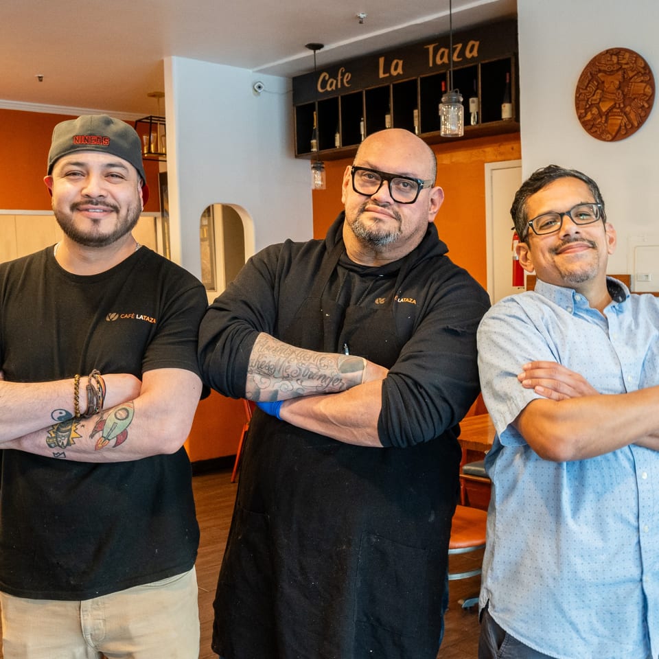 Three men, two wearing Cafe La Taza shirts, stand with crossed arms inside the cafe.