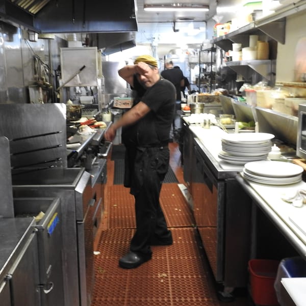 A chef in a yellow cap and black uniform stands in a busy commercial kitchen.
