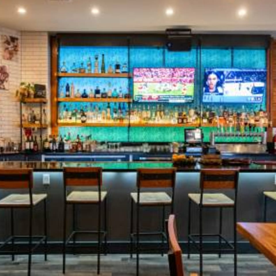 A modern bar area with liquor bottles on shelves, two TVs, and several bar stools.
