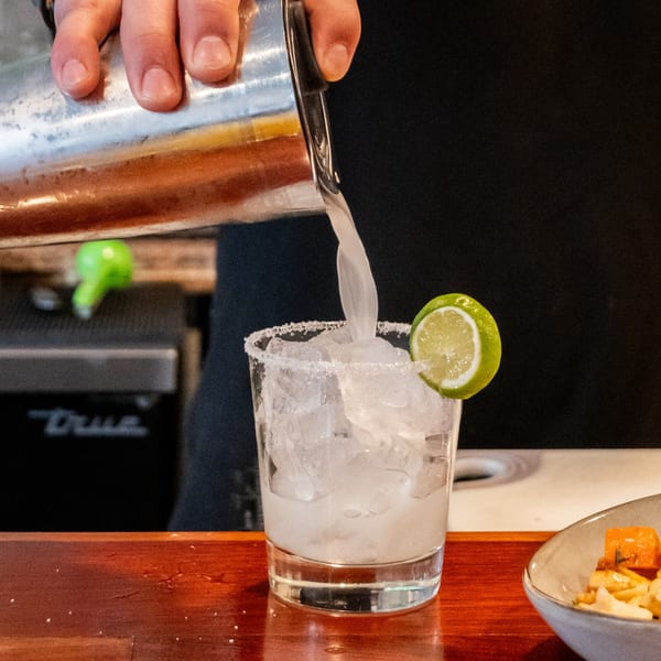 A bartender pours a margarita from a shaker into a salt-rimmed glass with ice and a lime.
