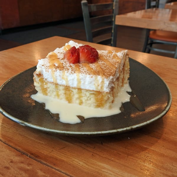 A slice of tres leches cake with whipped cream, strawberries, cinnamon, and caramel drizzle.