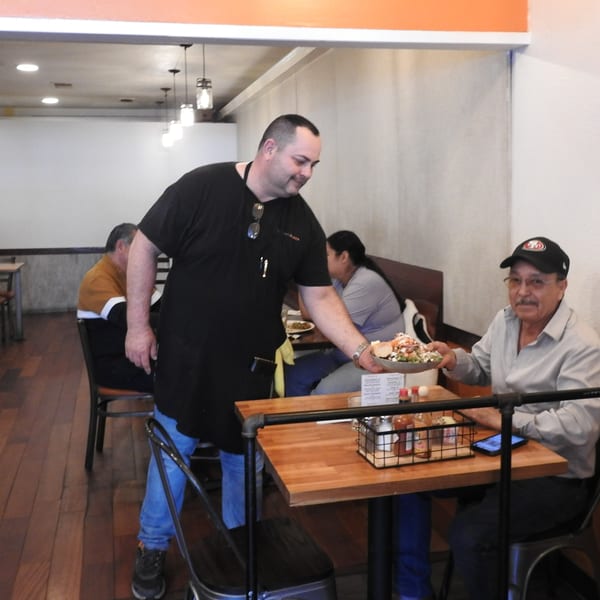A server in a black apron delivers a loaded plate of food to a smiling customer.