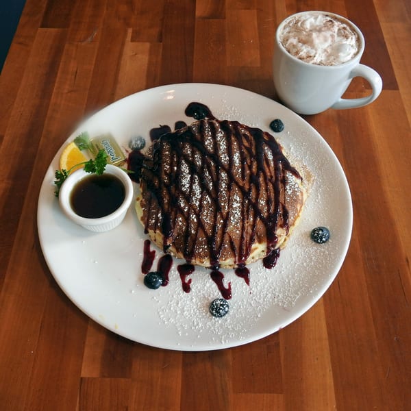 Chocolate drizzled pancakes with powdered sugar, blueberries, syrup, and a hot chocolate on a wooden table.